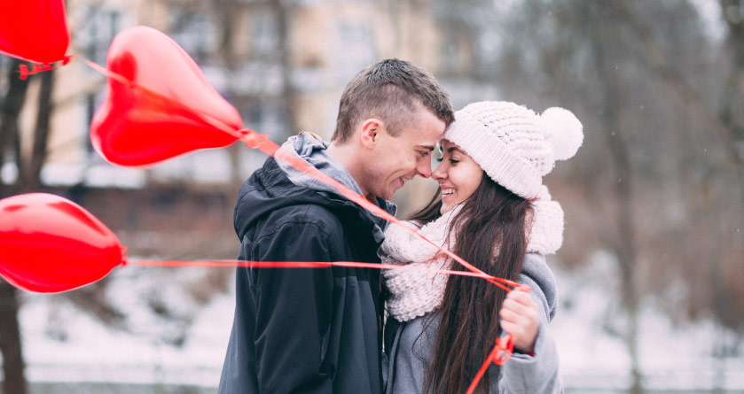 young man in black jacket and woman in winter hat and scarf standing outside in snow smiling at each other holding red heart balloons