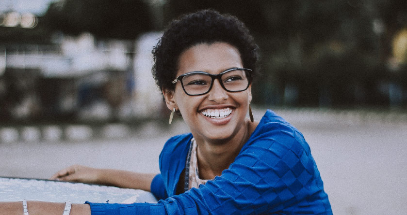 Close-up of a smiling dark-haired woman wearing black glasses and a blue shirt while leaning on a car