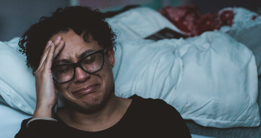 Closeup of a crying person with a headache due to sleep deprivation sitting on the floor next to their unmade bed