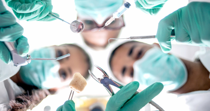 3 oral surgeons wearing mouth masks while holding surgery tools look down at a patient