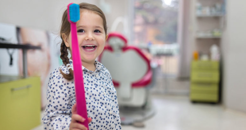 young girl at the dentist's office holds up a giant toothbrush after learning about good oral hygiene