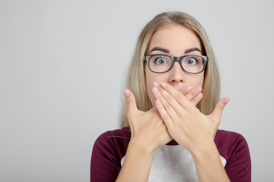 young woman wearing glasses covers her mouth with both hands to hide her gums
