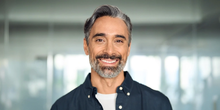 Middle-aged man smiling at camera in bright office setting wearing dark button-up shirt.